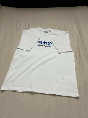 Vit Maison Margiela t-shirt - Vit t-shirt från Maison Margiela med blå logga och text på bröstet. Klassisk rak passform och rund halsringning. Tillverkad i mjuk bomull som känns skön mot huden. Perfekt för dig som gillar stilrena och trendiga plagg.