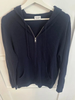 Mörkblå hoodie från John Henric - Mysig mörkblå hoodie från John Henric med dragkedja framtill, två fickor och justerbar huva med snören. Tillverkad i ett mjukt material som känns skönt mot huden. Perfekt för en avslappnad och stilren look. Nypris 2999kr