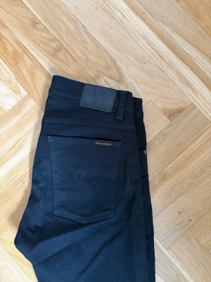 Svarta slim jeans från Nudie Jeans - Säljer ett par svarta slim fit jeans från Nudie Jeans. Klassisk femficksmodell med smal passform och stretchigt material för extra komfort. Snygga detaljer som svarta sömmar och diskret logga på bakfickan. Perfekta till en avslappnad streetwear-look.