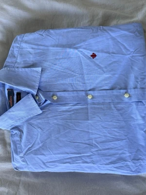 Blå skjorta från Morris Stockholm - Klassisk ljusblå skjorta från Morris Stockholm med broderad logga på bröstet. Skjortan har knappar framtill och en stilren krage. Tillverkad i mjuk bomull som känns skön mot huden. Perfekt för dig som gillar en clean och tidlös look.