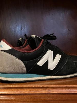 Svarta New Balance sneakers - Snygga svarta New Balance sneakers med vit logga på sidan, grå mockadetalj vid hälen och vinröda inslag runt öppningen. Skorna har en blå rand på mellansulan och klassisk låg siluett. Perfekta för dig som gillar retrostil och bekväma material.