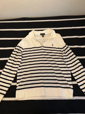 Randig tröja från Polo Ralph Lauren - Vit långärmad tröja med mörkblå horisontella ränder från Polo Ralph Lauren. Tröjan har en klassisk krage med dragkedja och den ikoniska röda logotypen broderad på bröstet. Tillverkad i mjukt bomullsmaterial för en skön känsla. (Barnstorlek)