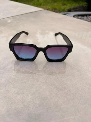 Svarta solglasögon med färgade glas - Säljer ett par trendiga solglasögon med svart, kantig båge och glas som skiftar i blått och rosa. Snygg statement-accessoar med cool retrovibe och breda skalmar. Perfekta för att lyfta vilken outfit som helst.🕶️☀️⛱️