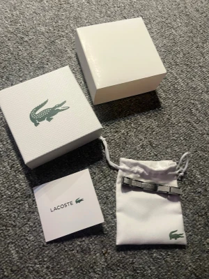 Silverfärgad klocka från Lacoste - Armband från Lacoste i silverfärgat metallarmband. Kommer med original ask och tygpåse med Lacoste-logga. Perfekt accessoar för dig som gillar klassisk och enkel design.
