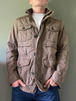 Beige fieldjacket - Snygg beige fieldjacket med flera stora fickor framtill och hög krage. Jackan har knappar och dragkedja samt lång ärm. Jackan är från märket clique.