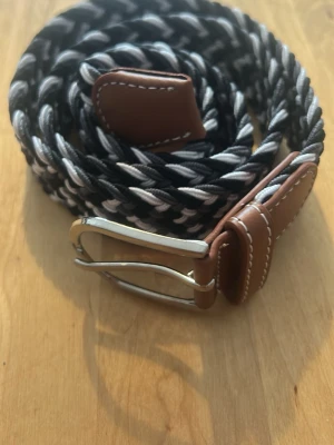 Flätat svartvitt bälte med läderdetaljer - Snyggt flätat bälte i svart och vitt med bruna läderdetaljer och silverfärgat spänne. Perfekt accessoar för att lyfta din outfit med en cool kontrast. Bältet har vita sömmar på läderdelarna och en klassisk metallspänne.