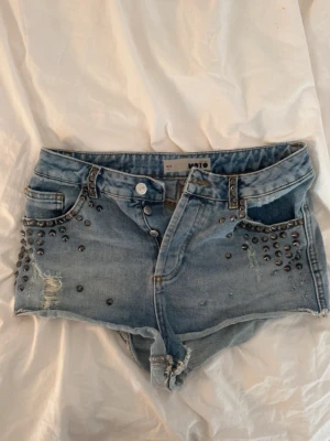 Blå jeansshorts med nitar från Moto - Säljer ett par ljusblå jeansshorts från Moto med coola nitar och slitningar framtill. Shortsen är högmidjade och har rå kant nedtill för en edgy look. Perfekta för sommaren och festivaler!