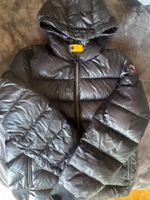 Svart dunjacka från Parajumpers - Snygg svart dunjacka från Parajumpers med huva och dragkedja framtill. Jackan har ett gult spänne vid halsen och klassisk Parajumpers-logga på ärmen. Tillverkad i glansigt material och fylld med dun för extra värme. Perfekt för kalla vinterdagar.