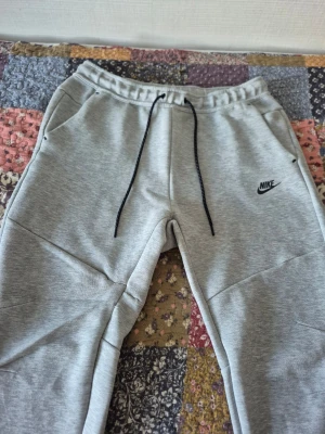 Nike tech fleece byxor  - Snygga gråa mjukisbyxor från Nike med svart logga på vänster ben. Byxorna har resår i midjan med svarta dragsnören och två sidofickor. Tillverkade i mjukt sweatshirtmaterial som är perfekt för chill eller träning. På höger sida fanns det hål men lyckades att sy ihop det och det är inte så synlig. 🙏🏼 