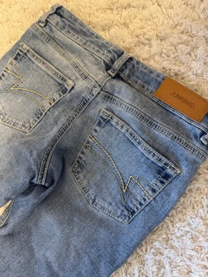 Ljusblå jeans från Junkyard - Säljer ett par ljusblå jeans från Junkyard med klassisk femficksdesign och coola kontrastsömmar på bakfickorna. Jeansen har en rak passform med utsvängda ben (bootcut/flair) och är tillverkade i denim med snygg tvätt. Perfekta för en avslappnad och trendig look.