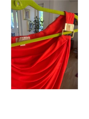 Röd oneshoulder klänning Michael Kors - Röd oneshoulder klänning från Michael Kors med snygg drapering och guldfärgad detalj vid axeln. Tillverkad i ett mjukt och stretchigt material som ger en smickrande siluett.