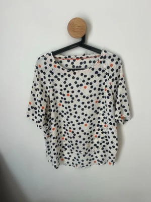 Prickig topp från Lindex  (Holly & Whyte) - Snygg topp från Holly & Whyte by Lindex med vitt tyg och prickigt mönster i marinblått och orange. Modellen har rund halsringning och korta ärmar. Perfekt för dig som gillar lekfulla prints och en avslappnad stil.
