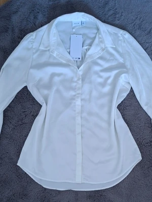Vit långärmad blus från Vila - Klassisk vit blus från Vila med lång ärm och bred krage. Blusen har en figurnära passform och knäppning framtill. Tillverkad i ett mjukt, glansigt material som ger en elegant look. Perfekt att styla med jeans eller kjol.