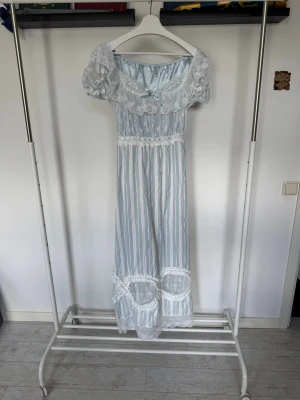 Långrandig blåvit klänning från Lacemade! - Romantisk långklänning i blått och vitt med vertikala ränder och breda spetsdetaljer vid halsringning, midja och nederkant. Klänningen kan knytas i ryggen för att framhäva midjan! Här kommer måtten från LaceMades hemsida! Bröst: 109cm, Midja: 98cm