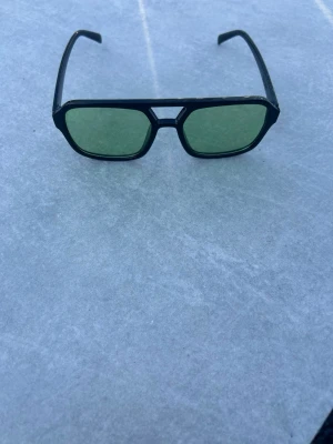 Svarta retro solglasögon med gröna glas - Snygga solglasögon med svart, kantig båge och gröna glas. Modellen har en cool retrovibe med breda skalmar och unik detalj vid näsryggen. Perfekta för dig som vill sticka ut med en klassisk men edgy stil. Väldigt fina och perfekta till sommaren och gött häng med polarna 