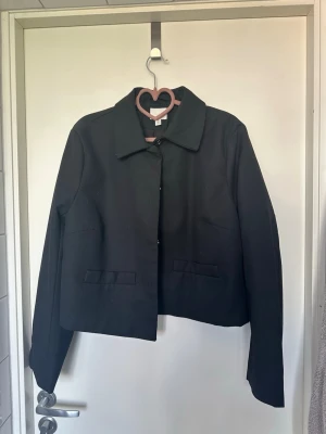 Svart jacka från H&M med klassisk krage - Stilren svart jacka/ kort kappa/kavaj från H&M med klassisk krage och raka linjer. Jackan har två fickor framtill och stängs med knappar. Perfekt för dig som gillar enkel och tidlös design. Passar till många olika outfits.