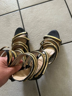 Svarta sandaler med gulddetaljer storlek 38 - Snygga svarta sandaler med flera breda remmar i guld och svart. Sandalerna har öppen tå och dragkedja bak för enkel påtagning. Perfekta för dig som vill sticka ut med en trendig och glamorös look. Materialet ser ut att vara konstläder.