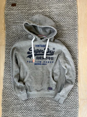 2000s vintage Superdry hoodie - Säljer en grå hoodie från Superdry med stort blått tryck på bröstet och japanska tecken. Klassisk känguruficka framtill, vita dragsnören i huvan och ribbade muddar. Tillverkad i mjuk bomullsblandning för skön känsla. Perfekt för dig som gillar streetwear och snygga detaljer.