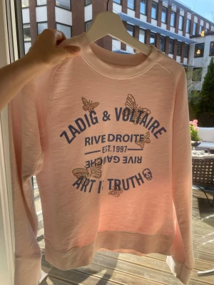 Rosa sweatshirt Zadig & Voltaire - Ljusrosa sweatshirt från Zadig & Voltaire med blå texttryck och glittriga fjärilar framtill. Rund halsringning, långa ärmar och mjukt material. Snygg detalj med döskalle och texten 'Rive Droite' och 'Art is Truth'. Perfekt för en chill och trendig look.
