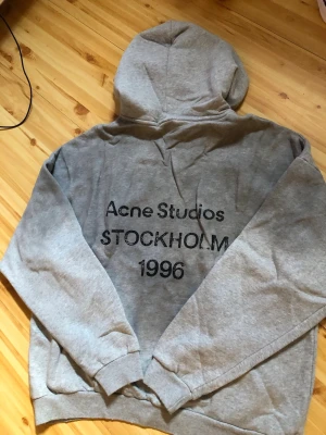 Grå hoodie Acne Studios Stockholm - Säljer en grå hoodie från Acne Studios med trycket 'Acne Studios STOCKHOLM 1996' på ryggen. Klassisk passform med huva och ribbade muddar. Perfekt för dig som gillar streetwear och vill ha en stilren look.