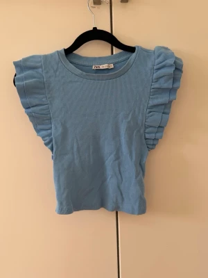 Blå ribbad topp med volang från Zara - Supersöt ljusblå ribbad topp från Zara med volangdetaljer längs axlarna. Toppen är ärmlös och har en croppad passform som ger en trendig look. Perfekt att styla med jeans eller kjol för en fräsch outfit.