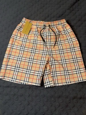 Rutig Burberry shorts - Snygga shorts från Burberry i klassiskt beige rutigt mönster med inslag av svart, vitt och rött. Modellen har elastisk midja med svart snörning och är tillverkad i ett lätt och bekvämt material. Perfekta för sommaren och ger en trendig look. PASSAR M. HELT NY, OANVÄND.