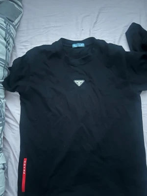 Svart t-shirt från Prada - Snygg svart t-shirt från Prada med klassisk rund hals och ikonisk trekantig logga på bröstet. T-shirten har korta ärmar och en röd detalj med Prada-tryck på sidan. Perfekt för dig som gillar stilrena designer och exklusiva märken.