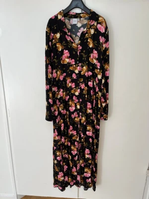 Blommig långklänning från ICHI - Långklänning från ICHI med svart bas och stora blommor i rosa, brunt och vitt. Klänningen har långa ärmar, knappar framtill och en klassisk krage. Perfekt för dig som gillar färgstarka mönster och en avslappnad men ändå stilsäker look.