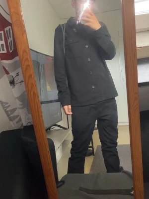 Svart overshirt med knappar - Stilren svart overshirt med klassisk krage och knäppning framtill. Jackan har raka linjer, två bröstfickor och är långärmad. Perfekt för lager-på-lager och enkel att matcha med olika outfits.