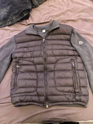 Grå dunväst från Moncler - Snygg grå dunväst från Moncler med stickade ärmar och rygg i mjukt material. Västen har två dragkedjeförsedda fickor fram, hög krage och Moncler-logga på ärmen. Perfekt för lager-på-lager och ger en sportig look.