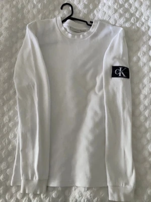 Vit långärmad tröja Calvin Klein - Vit långärmad tröja från Calvin Klein med rund halsringning och svart CK-logga på vänster ärm. Tröjan har ribbade muddar vid ärmslut och är gjord i ett mjukt, bekvämt material. Perfekt för en clean och stilren look.
