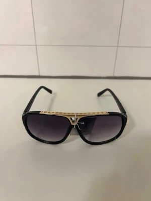 Louis Vuitton svarta solglasögon - Säljer ett par snygga svarta solglasögon från Louis Vuitton med guldiga detaljer på ovansidan av bågen och LV-logga. Glasen är mörka och har en klassisk fyrkantig form. Perfekt för dig som vill sticka ut med exklusiva accessoarer.