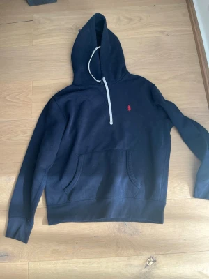 Mörkblå hoodie från Polo Ralph Lauren - Snygg mörkblå hoodie från Polo Ralph Lauren med klassisk röd logga broderad på bröstet. Hoodien har huva med vita dragsnören och en stor magficka framtill. Tillverkad i mjukt bomullsmaterial som känns skönt mot huden. Perfekt för en avslappnad och stilren look.