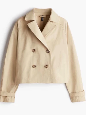 Beige kort trenchcoat från H&M - Stilren beige trenchcoat från H&M med dubbelknäppning och bred krage. Kortare modell med långa ärmar och dekorativa knappar vid ärmsluten. Tillverkad i ett kraftigare bomullstyg som ger en klassisk look. Tror det är maskara på handleden, har ej försökt ta bort