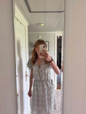 Vit blommig kortklänning med volang - Söt vit kortklänning med små blommor i svart och beige. Klänningen har v-ringning, volangärmar och ett knytband i midjan för en feminin siluett. Perfekt för varma dagar och har en luftig passform.