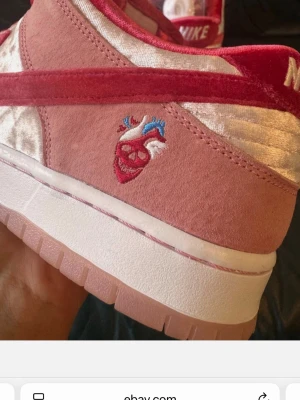 Nike SB Dunk Low StrangeLove rosa - Nike SB Dunk Low StrangeLove i rosa och rött med sammetsdetaljer och broderat hjärta på sidan. Skon har en vit och rosa sula, röda skosnören och klassisk Nike-logga på hälen. Materialmix av mocka och sammet ger en unik look.