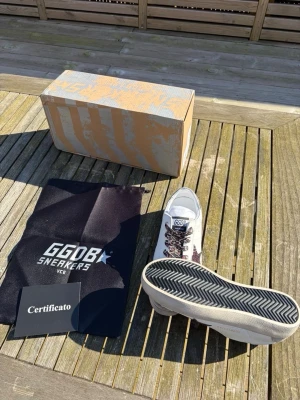 GGDB vita sneakers med glitterdetaljer - Snygga vita GGDB sneakers med beige mockadetaljer på tån och en glittrig mörklila stjärna på sidan. Skorna har leopardmönstrade skosnören och klassisk låg silhuett. Tillverkade i läder och mocka, levereras med originalkartong, dustbag och certifikat.