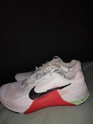 Nike Metcon 7 vita och röda sneakers - Nike Metcon 7 sneakers i vitt meshmaterial med svarta swoosh-loggor och röda samt mintgröna detaljer på sulan. Skorna har en sportig look med låg profil och snörning. Perfekta för dig som gillar träning och streetstyle.