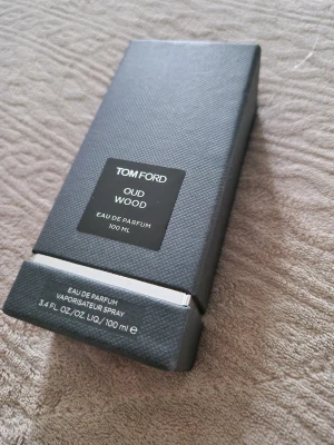Tom Ford Oud Wood Eau de Parfum 100ml - Tom Ford Oud Wood Eau de Parfum 100ml i elegant, mörkgrå rektangulär flaska med matchande lyxig kartong. Flaskan har en svart etikett med vit text och silverdetaljer. Doften är känd för sina träiga och exklusiva noter.