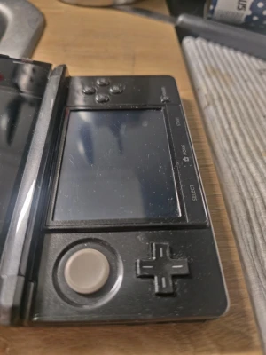 Nintendo 3DS svart bärbar konsol - Säljer en svart Nintendo 3DS, en bärbar spelkonsol med dubbla skärmar, styrspak, styrkors och klassiska knappar. Konsolen har en vikbar design och är tillverkad i plast med en blank finish. Perfekt för dig som gillar att spela på språng.
