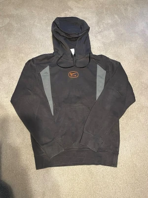Nike hoodie  - Säljer en svart hoodie från Nike med grå paneler på sidorna och en Nike Air-logga i orange på bröstet. Hoodien har huva och en stor magficka. Perfekt för en sportig och avslappnad stil.