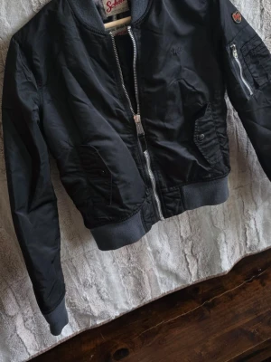 Svart bomberjacka från Schott NYC - Snygg svart bomberjacka från Schott NYC med klassisk ribbad mudd i ärmslut och midja. Jackan har dragkedja framtill, fickor med knapp och en extra ficka med dragkedja på ärmen. Perfekt för dig som gillar streetwear och tidlös stil.