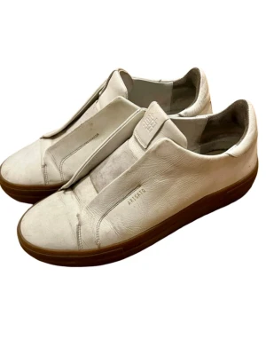 Vita sneakers från Axel Arigato - Stilrena vita sneakers från Axel Arigato med minimalistisk design och bruna gummisulor. Skorna är tillverkade i läder och har en slip-on look utan synliga snören. Märkeslogga på sidan och tungan. Perfekta för en clean och modern stil. I mycket bra skick skulle behöva en skovårdsrengöring bara. 