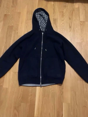 Mörkblå hoodie med mönstrat foder - Säljer en mörkblå hoodie med dragkedja och huva. Insidan har ett grått och svart mönstrat foder som ger en unik look. Tröjan har långa ärmar, två fickor framtill och justerbar dragsko i huvan.