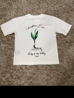 Vit Christian Dior t-shirt med tryck - Vit t-shirt från Christian Dior med korta ärmar och ett snyggt tryck på ryggen med texten 'Lily of the Valley 1947' och en blomillustration. Framtill finns en diskret Dior-logga. Tillverkad i mjuk bomull och har en klassisk passform.