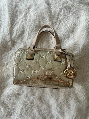 Guldig Michael Kors handväska - Snygg guldig handväska från Michael Kors med blank metallic finish och präglade MK-loggor över hela väskan. Väskan har två korta handtag i beige läder, guldfärgade metalldetaljer och en rund MK-tag. Invändigt finns ett fack och dragkedja upptill. Digitalt kvitto finns. 