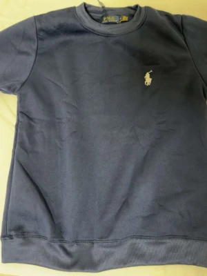 Blå crewneck från Polo Ralph Lauren - Mörkblå sweatshirt från Polo Ralph Lauren med klassisk rund halsringning och vit broderad logga på bröstet. Tröjan har långa ärmar och ribbade muddar vid ärmslut och nederkant. Perfekt för en chill och stilren look.
