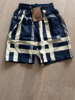 Burberry shorts - Snygga shorts från Burberry i klassiskt rutigt mönster med marinblå och beige färg. Shortsen har elastisk midja och sidofickor, samt Burberrys logga tryckt på ena benet. Perfekta för sommaren och ger en clean och trendig look. Inga defekter och i topp skick