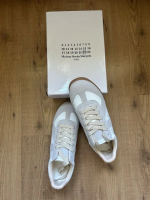 Maison Margiela vita sneakers - Snygga vita sneakers från Maison Margiela med beige mockadetaljer och vita läderpaneler. Klassisk låg modell med beige skosnören och gummisula. Perfekta för dig som gillar minimalistisk och stilren design.
