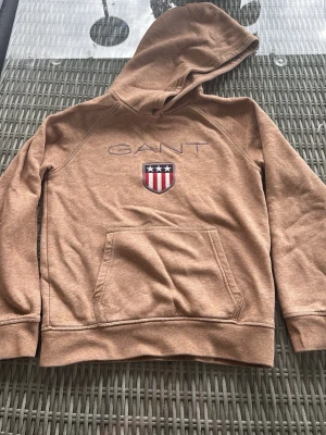 Brun hoodie från GANT  - Snygg brun hoodie från GANT med broderad logga på bröstet. Klassisk känguruficka framtill och varan är i nyskick. Stl 146/152